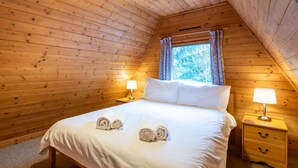 A cozy, wooden-paneled room with a neatly made bed, two bedside lamps, and a window with curtains. - Rowan Cottage by Interhome (Drumnadrochit)