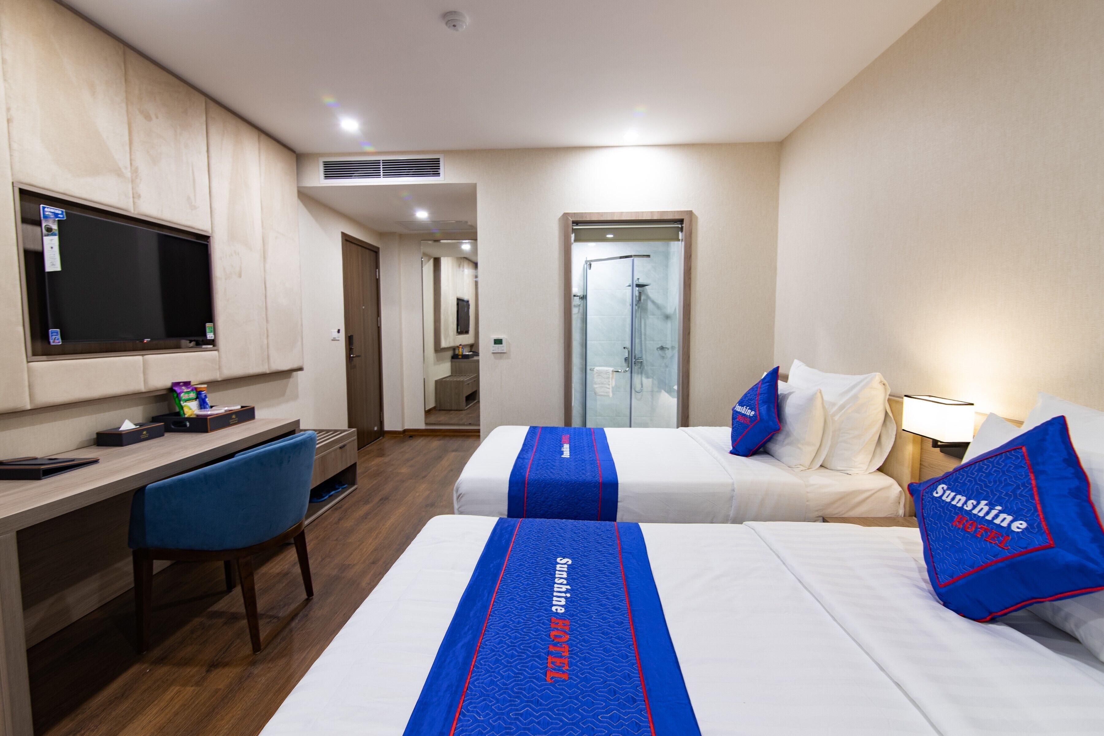 A hotel room with two beds, a desk, a chair, a TV, and a shower area.