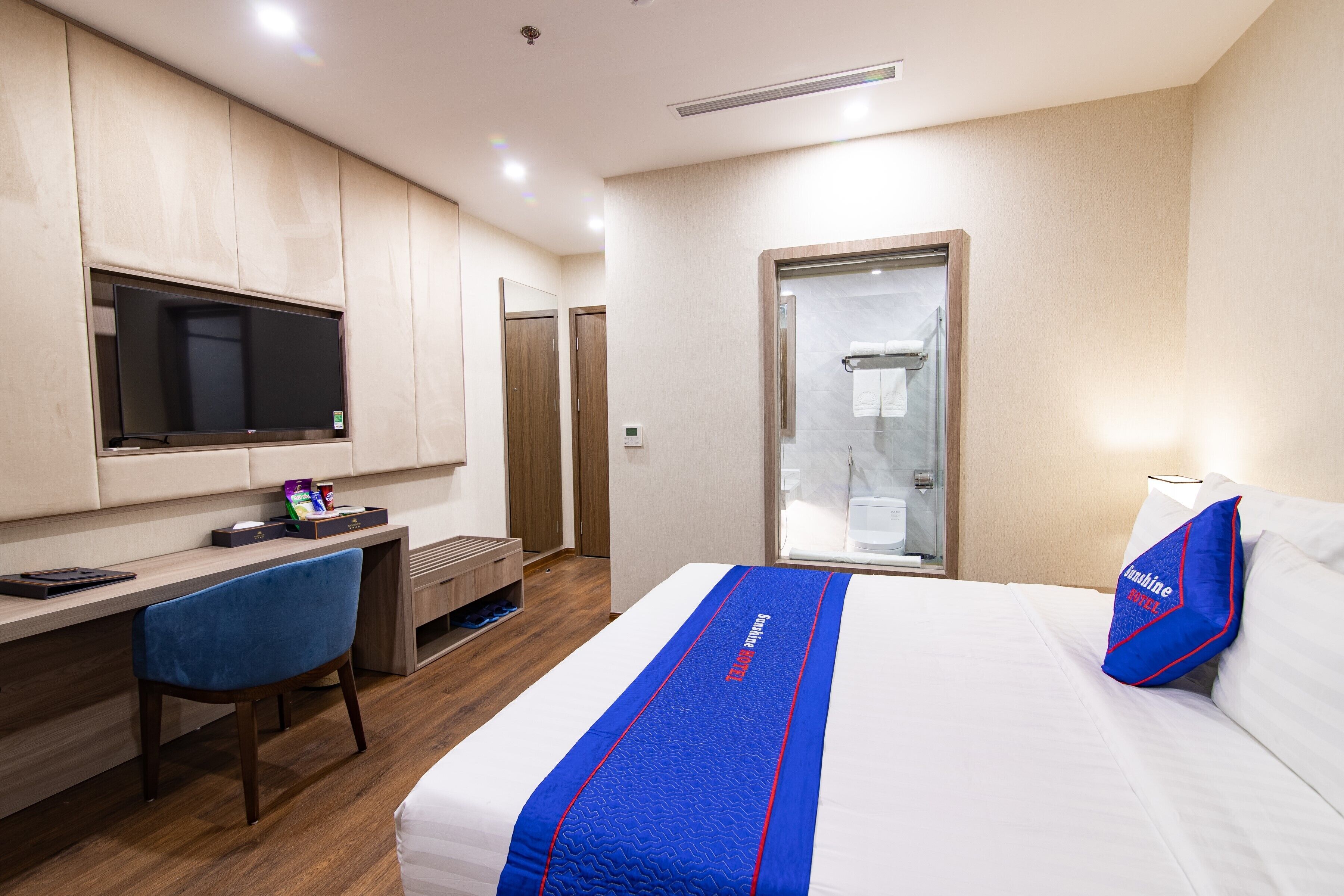 Habitación de hotel con cama, escritorio, silla, televisión y baño con puerta de cristal.