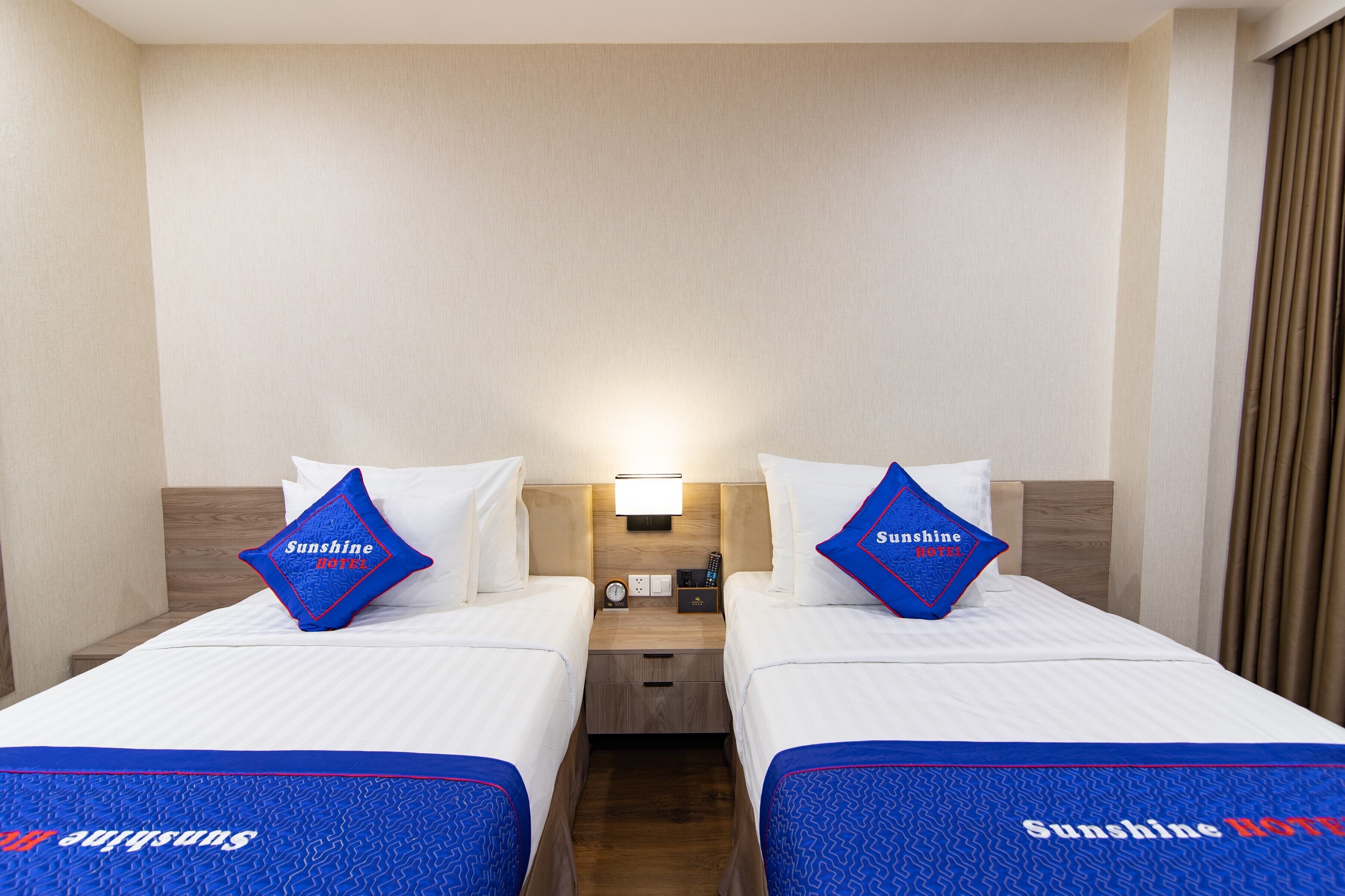 A hotel room with two beds, each with a blue bedspread and matching pillows, a nightstand with a lamp, and a wall-mounted clock.