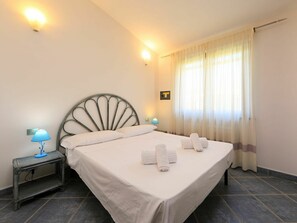 A neatly made bed with white linens and towels, a metal headboard, a bedside table with a lamp, and a window with sheer curtains.