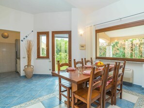A dining area with a wooden table and chairs, a vase with reeds, and a view of greenery through the window.