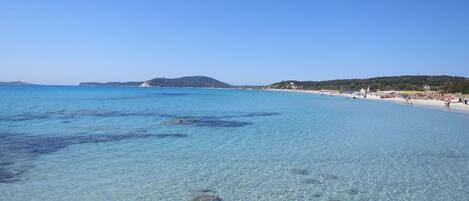 A clear coastal beach with rocky formations in the foreground and a distant hill.