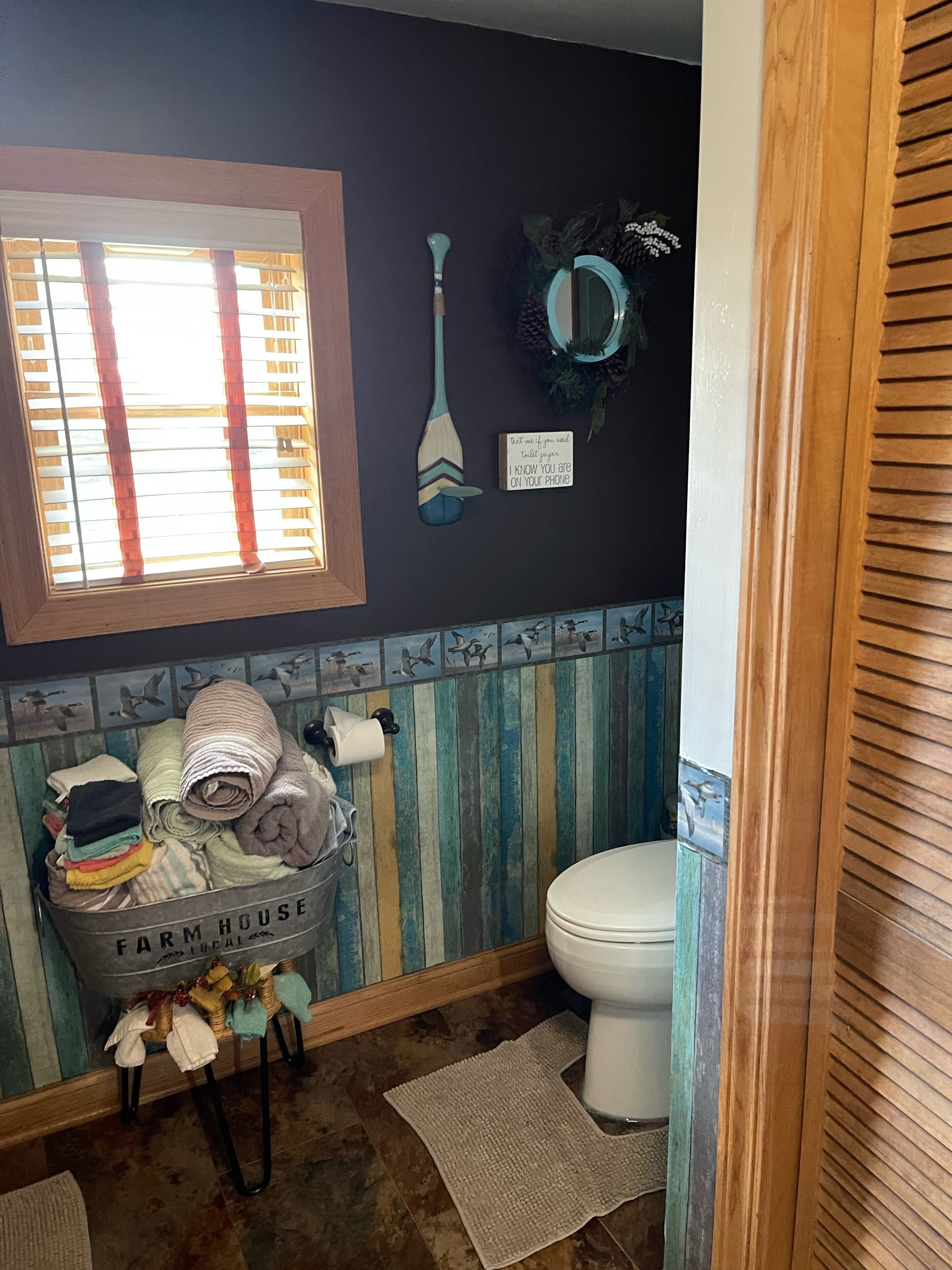 Une salle de bain avec des toilettes, une fenêtre avec des stores, un porte-serviettes fixé au mur et un panier étiqueté « FERME ».