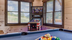 A pool table with billiard balls and a pool cue, a vintage arcade machine, and wooden walls with windows. - Modern Smoky Mtn Cabin, Hot Tub, Game Room, Pool Table, Near Dollywood (Sevierville)