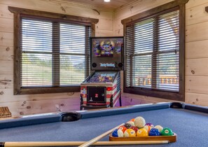 Ein Billardtisch mit Billardkugeln und Spielstange, ein alter Arcade-Automat und Holzwände mit Fenstern.