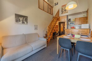 Een gezellige woonkamer met een bank, een eettafel gedekt voor twee personen, en een keuken met houten kastjes en een hangende lamp.