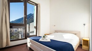 A hotel room with a bed, a window with curtains, a bedside table, and a view of mountains. - Cham Sud Balme401 Family 2BR Walk to Lifts & Centre, Chamonix, France (Chamonix)