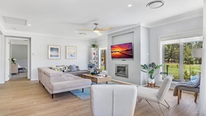 A modern living room with a sofa, coffee table, and a fireplace. There is a television mounted on the wall and a large window offering a view of the outdoors. - KOKOON - THE VINTAGE HOLIDAY HOUSES (POKOLBIN)