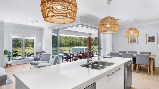 Eine moderne Küche mit einer großen Kochinsel, zwei hängenden Rattan-Kronleuchtern und Blick auf einen Pool und Grünflächen.