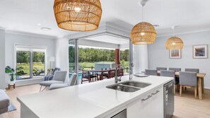 A modern kitchen with a large island, two hanging rattan chandeliers, and a view of a pool and greenery outside. - KOKOON - THE VINTAGE HOLIDAY HOUSES (POKOLBIN)