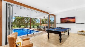 Uma área moderna de piscina interna com mesa de bilhar, uma espreguiçadeira e vista para a piscina e vegetação externa.