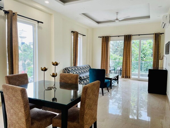 A spacious living area with a glass dining table, comfortable seating, and large windows offering a view of greenery. - Olive Service Apartments - DLF Cyber City (Gurugram)