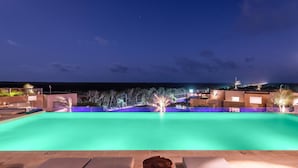 A nighttime view of a swimming pool with lounge chairs and a wooden table in the foreground, surrounded by buildings and a view of the sea. - Aloft Tulum (Tulum)
