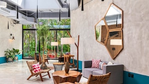 A modern lounge area with geometric wooden furniture, a large mirror, and a view of lush greenery through large windows. - Aloft Tulum (Tulum)