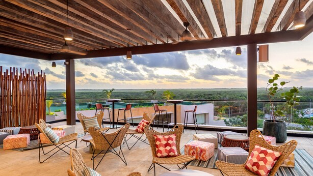 A covered outdoor seating area with wicker furniture and a view of a pool and landscape. - Aloft Tulum (Tulum)