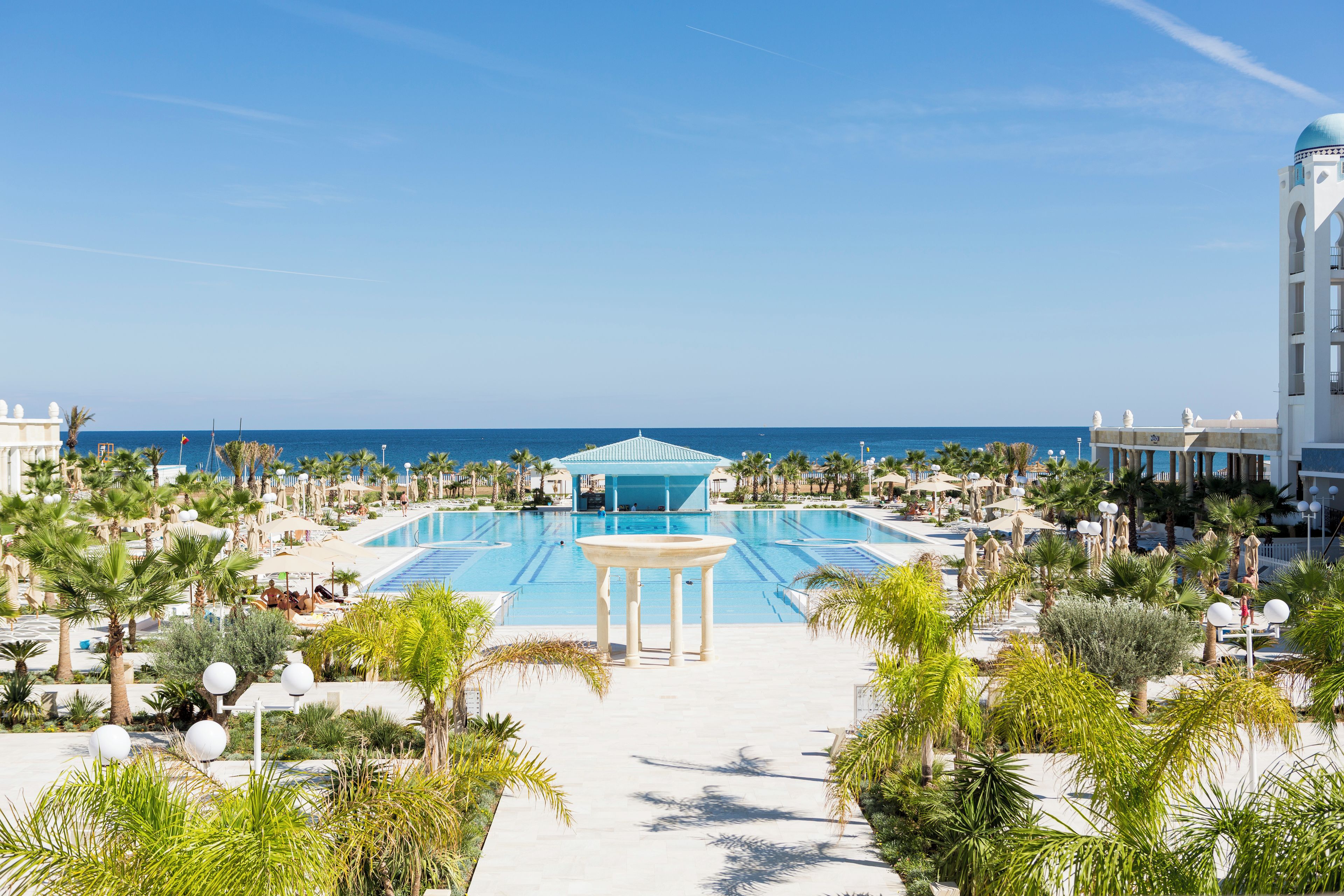 A resort pool area with a central pool, palm trees, and a view of the ocean.