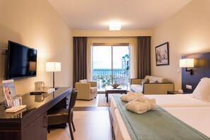 Een hotelkamer met een bed, een bureau met een televisie, een stoel, een bank, een balkon met uitzicht op zee, en een schilderij aan de muur.