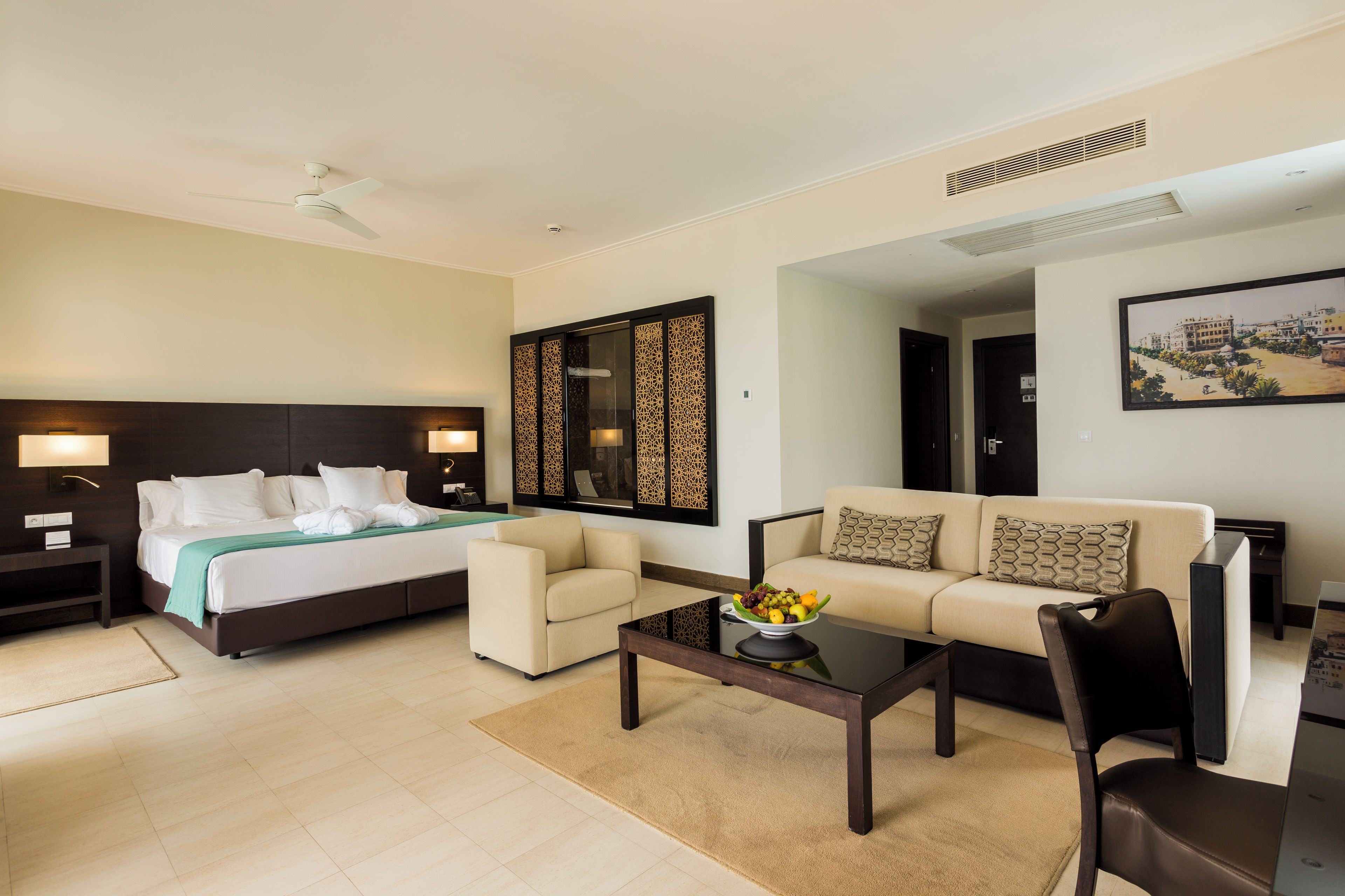 A hotel room with a bed, two sofas, a coffee table with fruit, and a painting on the wall.