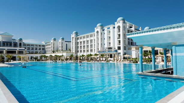 A large swimming pool with a clear blue water, surrounded by a hotel with multiple floors and a white facade. - Barcelo Concorde Green Park Palace (Port El Kantaoui)