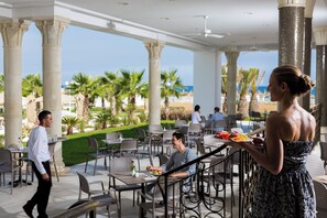 An outdoor dining area with tables and chairs, palm trees, and a view of the beach. - Barcelo Concorde Green Park Palace (Port El Kantaoui)