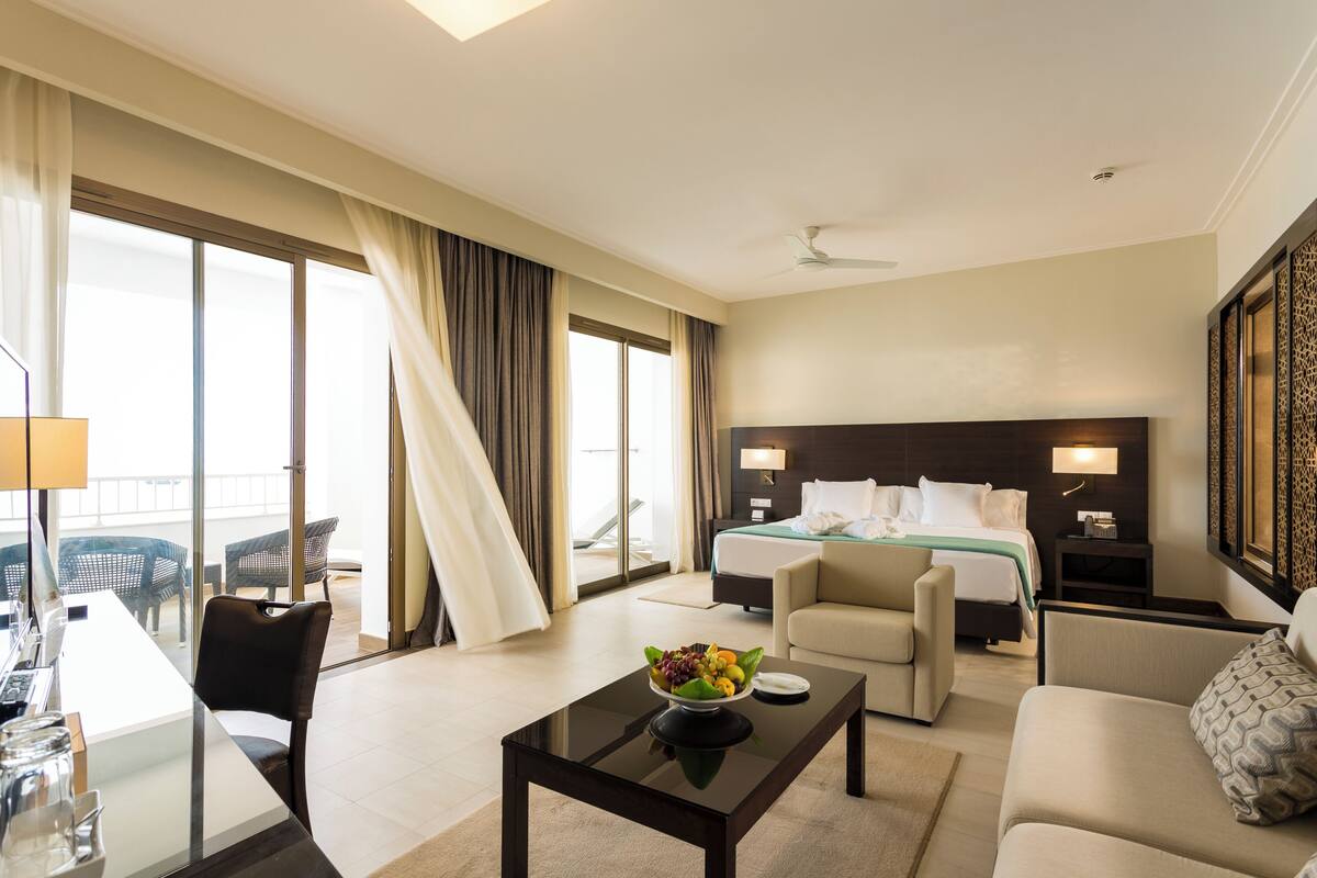 Habitación de hotel con cama, zona de estar con sofá y sillón, mesa de comedor con frutero, y ventana grande con cortinas.