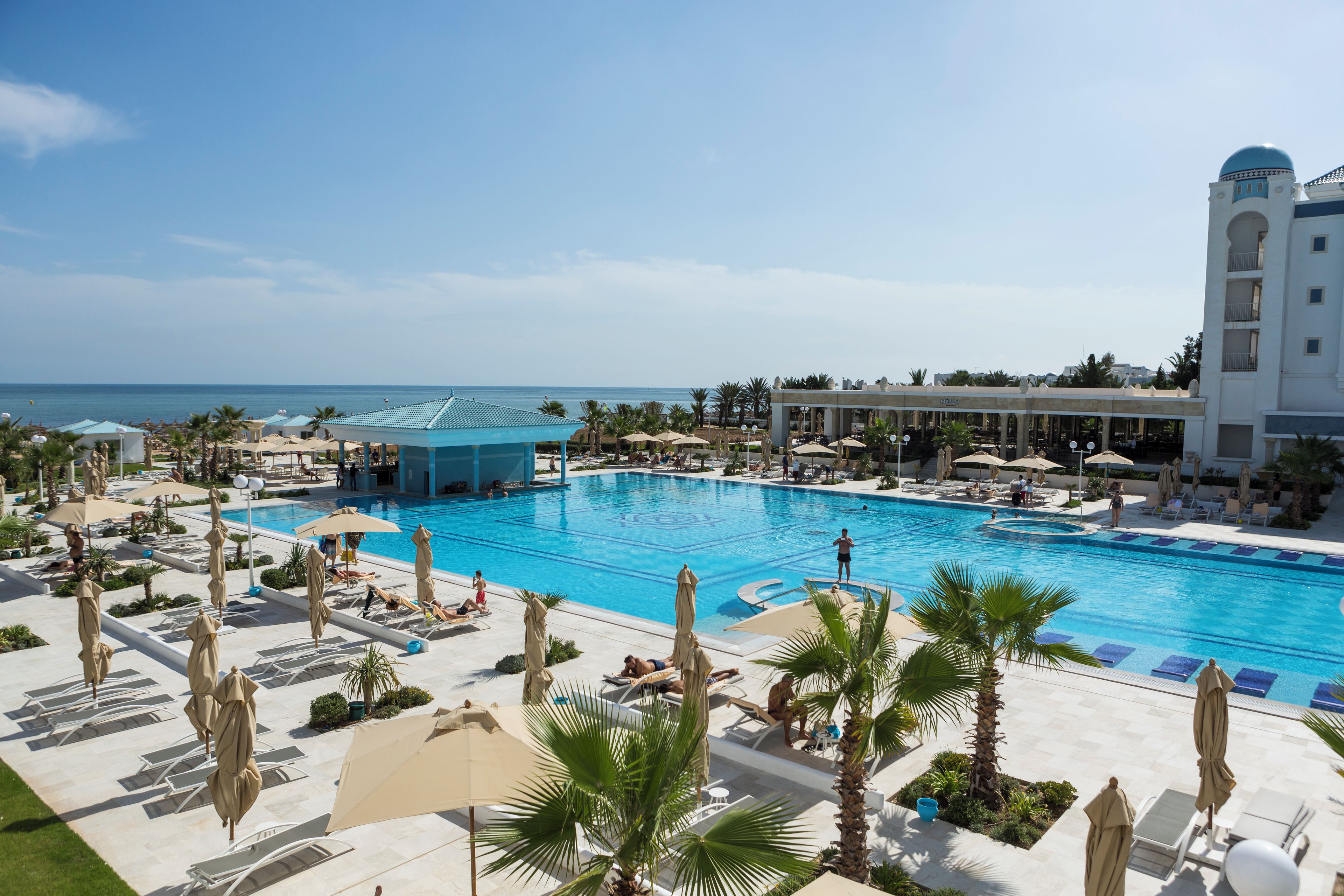 A large pool with multiple sun loungers and umbrellas, surrounded by palm trees and a building with a dome.
