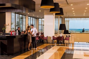 Un hall d’hôtel moderne doté d’un bar, de grandes fenêtres donnant sur la plage et d’un agencement varié des espaces de détente.