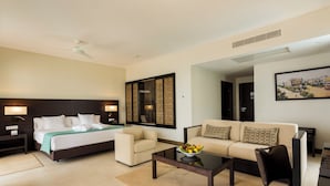 Habitación de hotel con cama, dos sofás, una mesa de centro con fruta y un cuadro en la pared.
