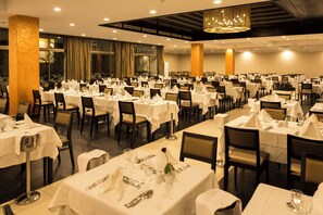Une grande salle à manger avec de nombreuses tables dressées, agrémentée d’un lustre moderne et de grandes fenêtres.