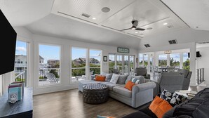 A spacious living room with a grey sectional sofa, a round ottoman, and a grey armchair. There are multiple windows offering a view of the neighborhood.