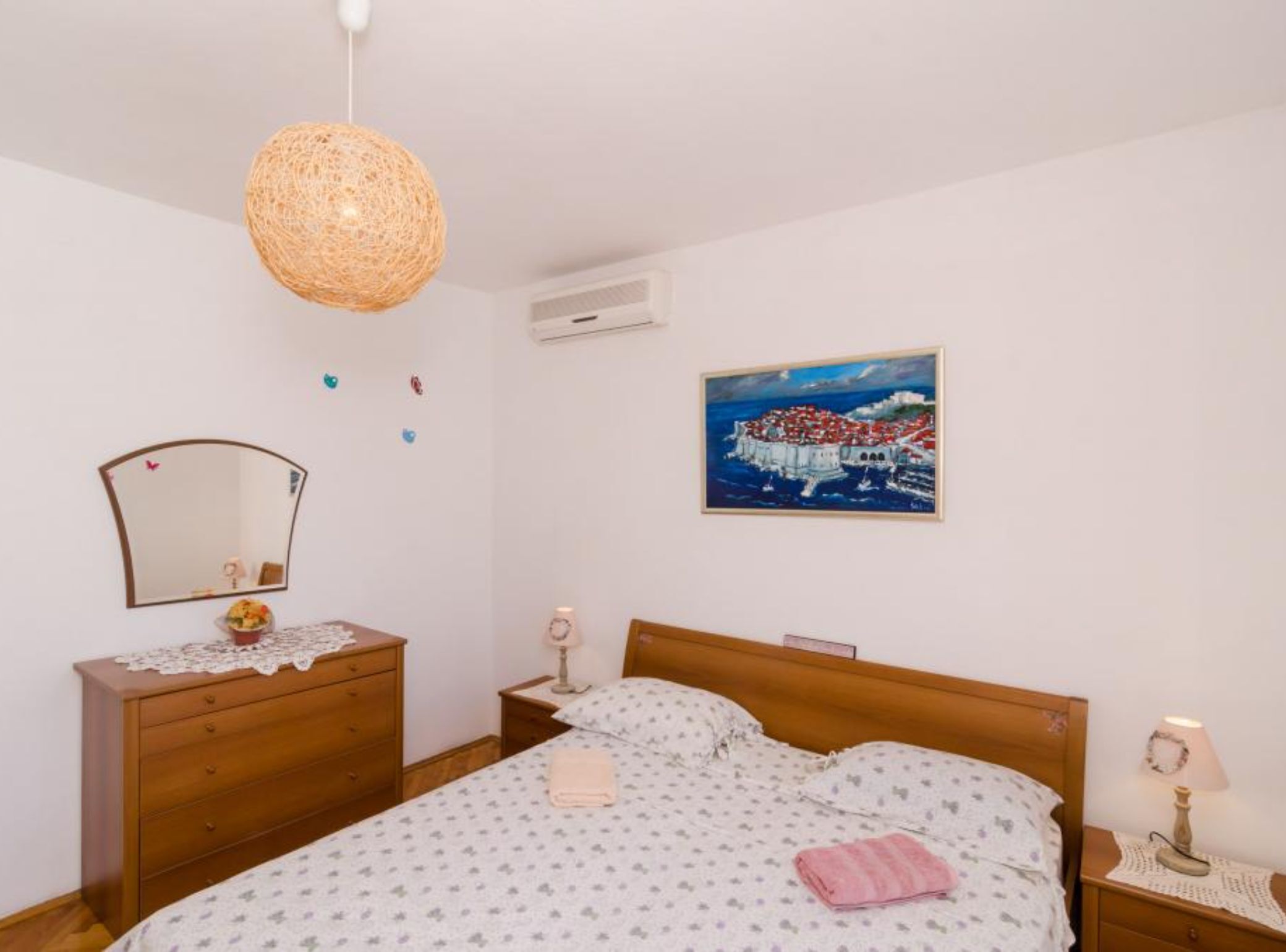Une chambre à coucher comprenant un lit, une commode en bois, une table de chevet, un miroir fixé au mur et un tableau représentant une ville côtière.