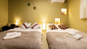 A hotel room with two beds, each with a pillow and a folded towel. There is an air conditioning unit on the wall, a nightstand with a lamp, and a small table with a chair. - Tokinosyukudaikokucyou 1F (Osaka)
