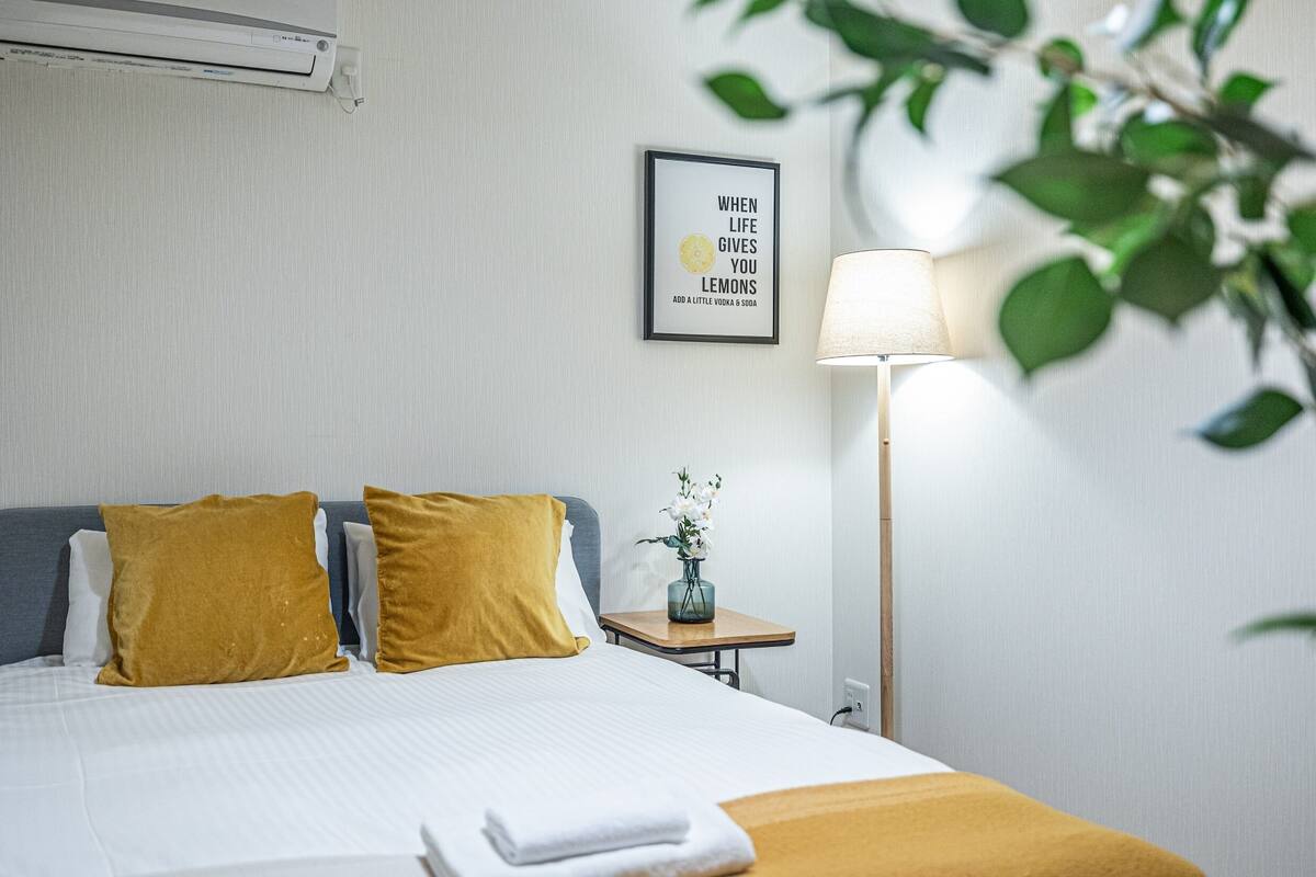Een keurig opgemaakt bed met twee kussens, een nachtkastje met een lamp, en een lijst met een citaat aan de muur.