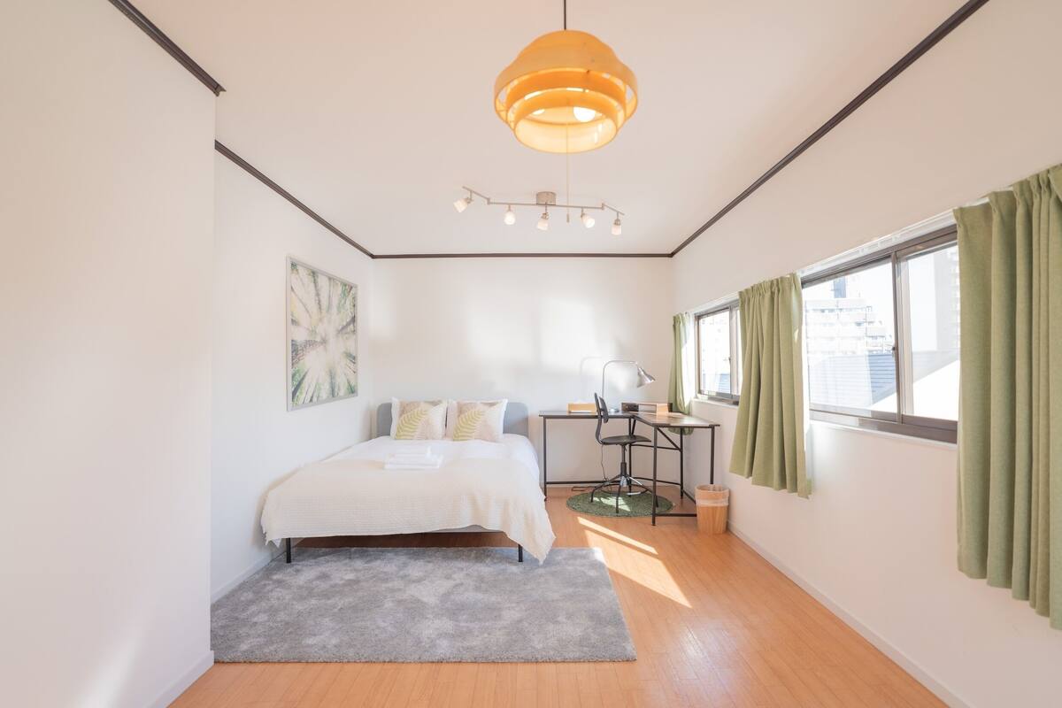 Een slaapkamer met een bed, bureau, stoel en een groot raam met gordijnen.