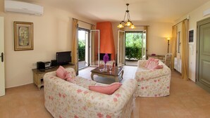 A living room with two floral sofas, a coffee table, and a TV. There is a fireplace and a view of greenery outside.