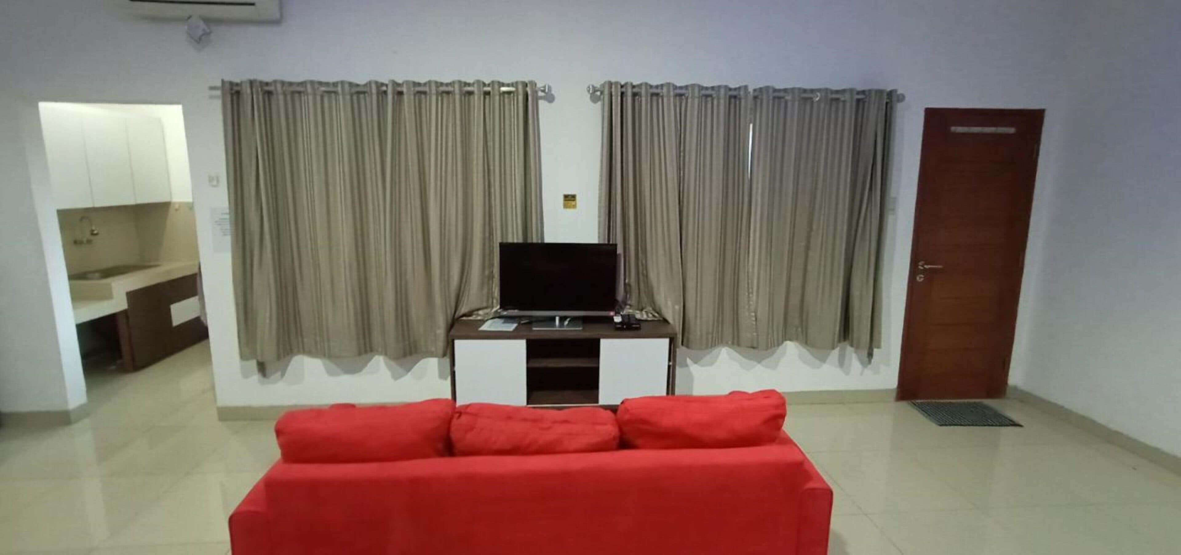 A living room with a red sofa, a TV on a white stand, and a kitchen area with a sink and cabinets.