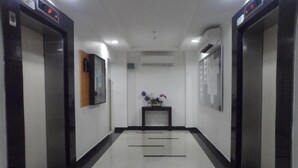 A hallway with two elevators, a wall-mounted mirror, a small table with a vase, and a bulletin board. - Sewaunitkalcit (Jakarta)
