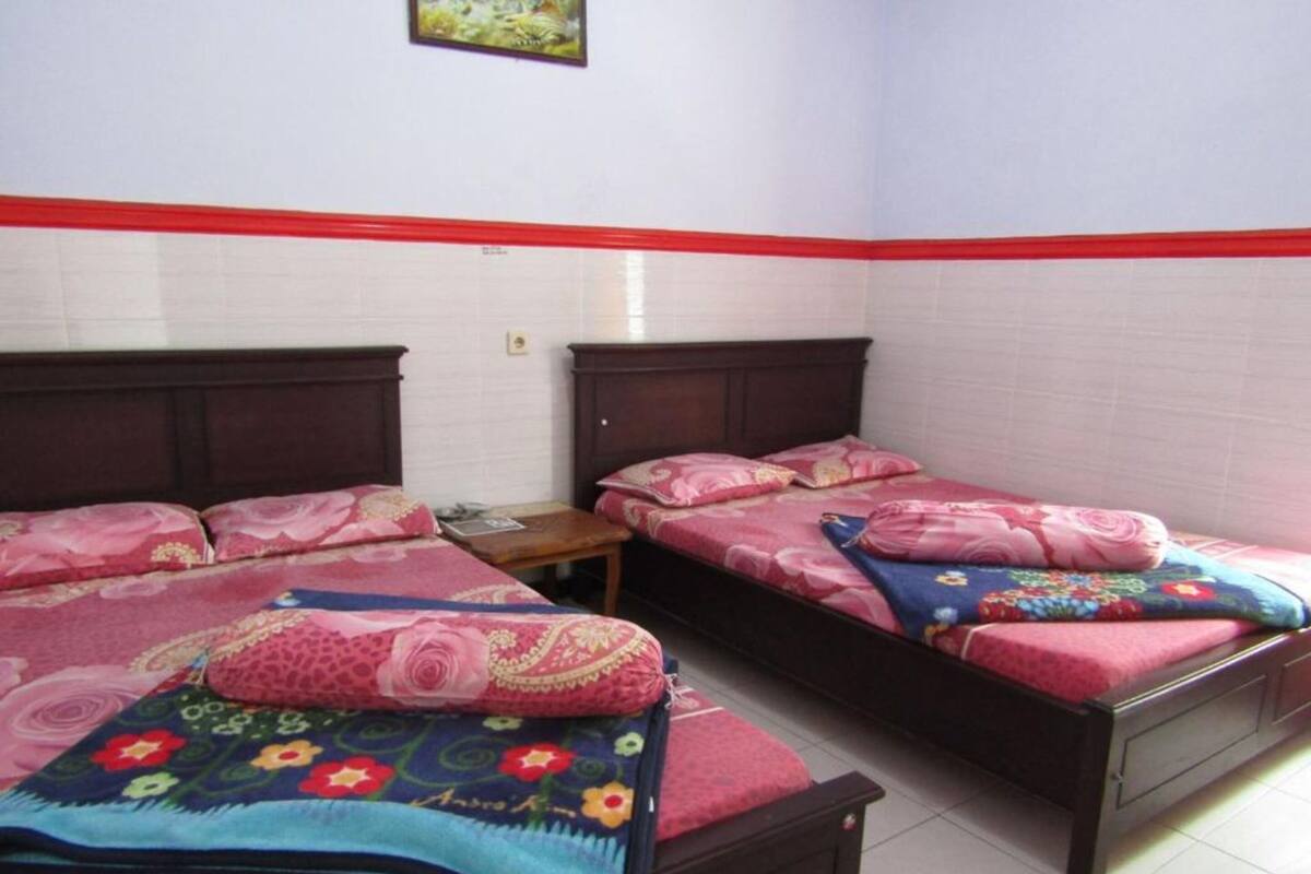 A room with two beds, each with floral bedding and wooden headboards. There is a small table between the beds, and a picture hangs on the wall above.