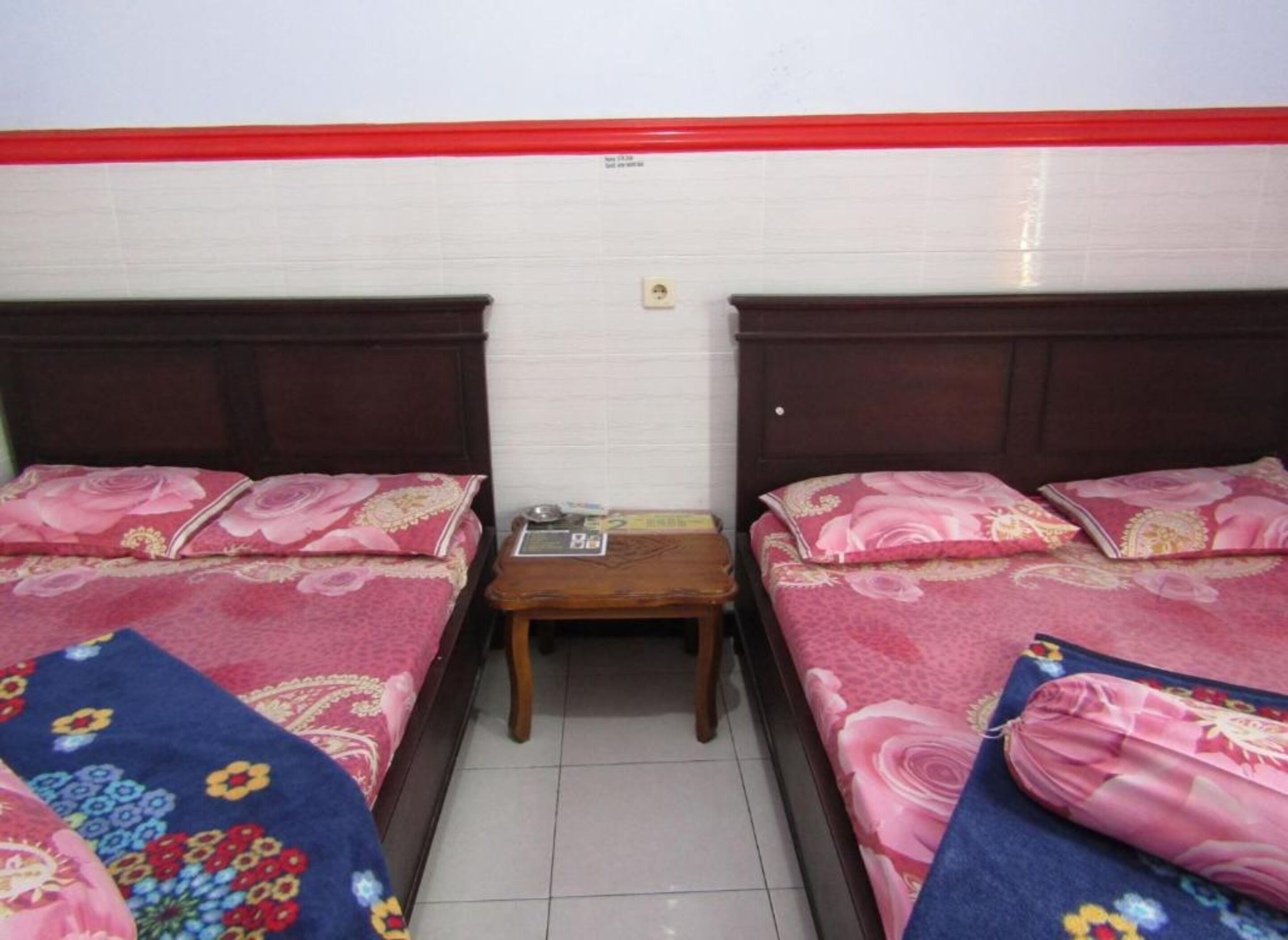 A room with two single beds, each with a floral bedspread and matching pillows. There is a small wooden nightstand between the beds with a few items on it. The room has white walls and a red trim at the top.