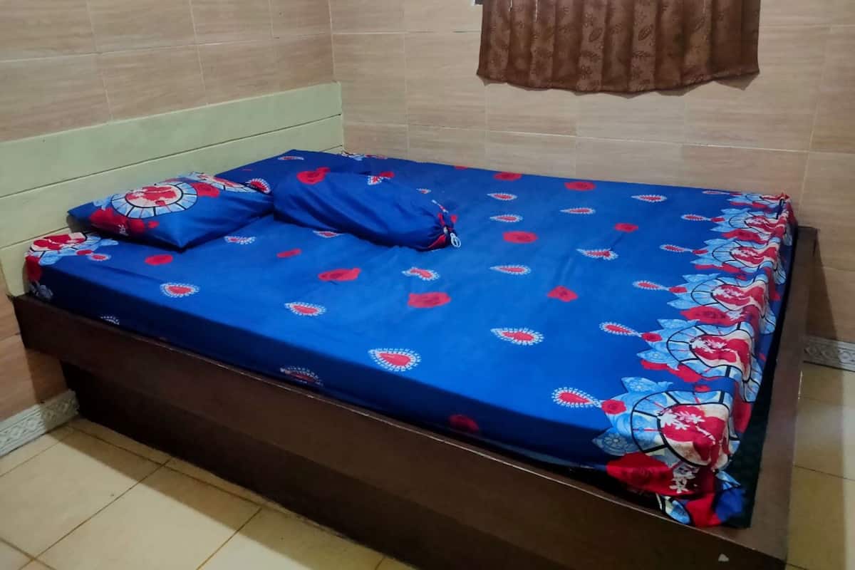 Un lit recouvert d’une couvre-lit bleue ornée de motifs floraux rouges et blancs, avec une tête de lit en bois et un rideau marron.