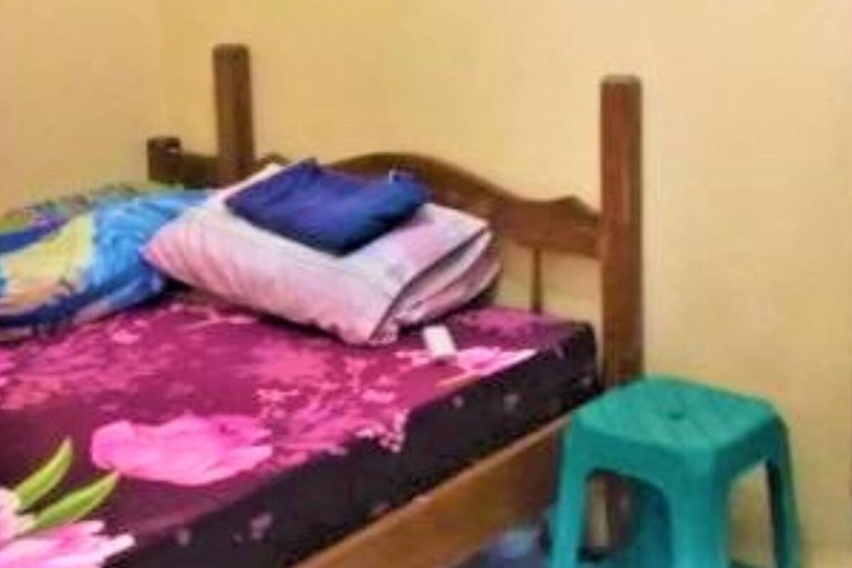 Una habitación pequeña con cama, cabecera de madera, un taburete color turquesa y una colcha de flores rosas.