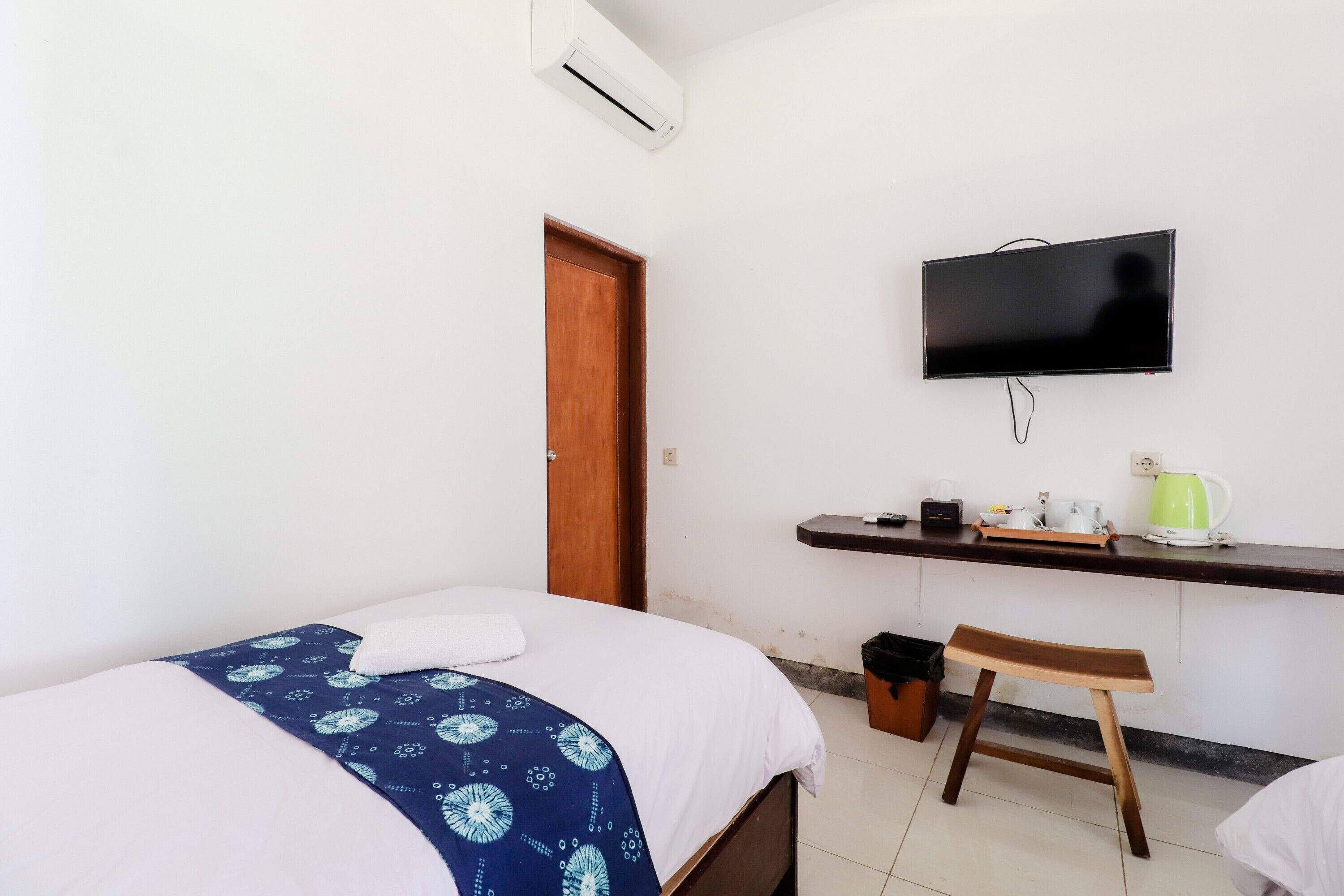 A bedroom with a bed, a wooden door, a television mounted on the wall, a small stool, and a shelf with a kettle and cups.