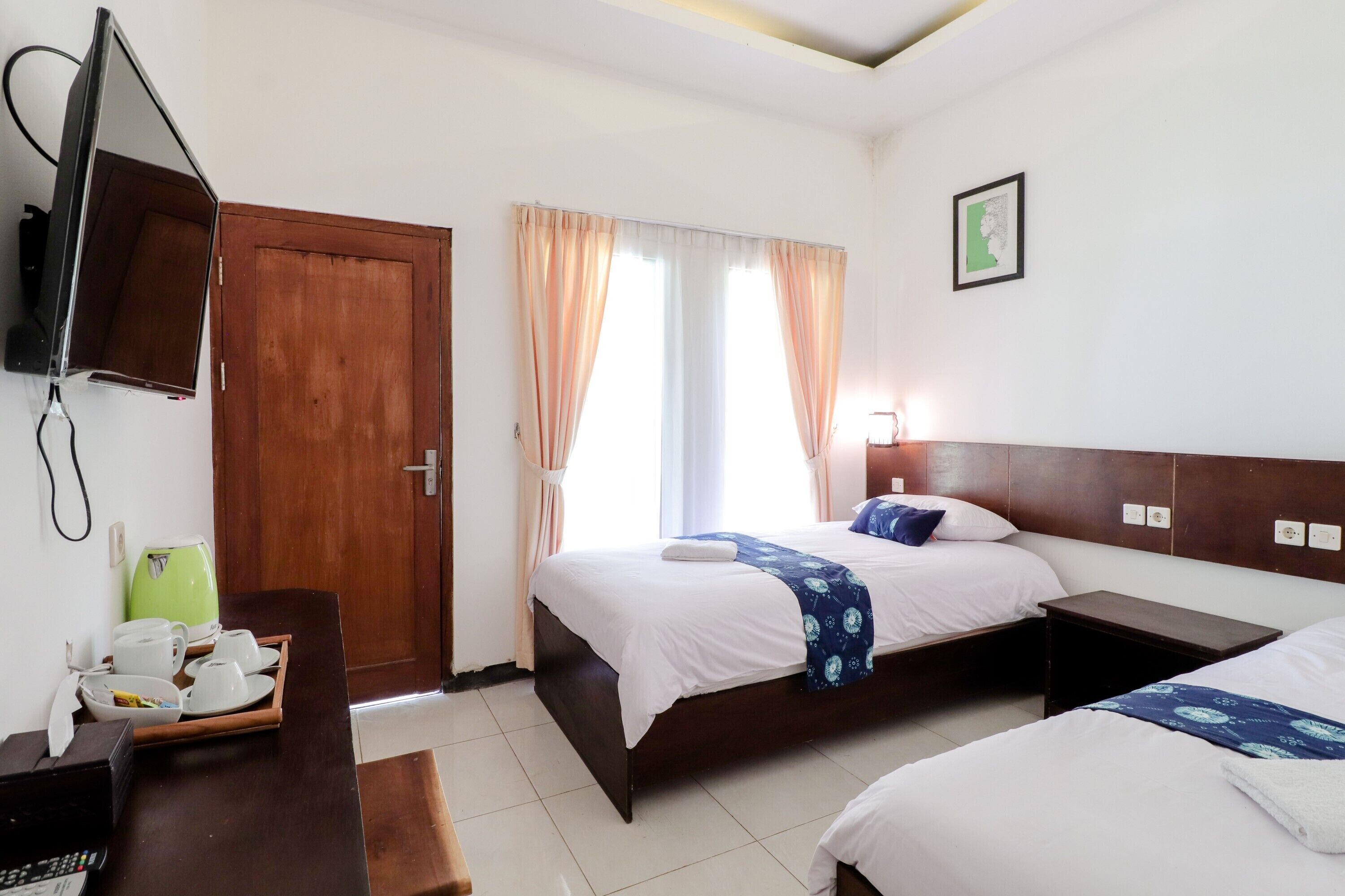Une chambre d’hôtel avec deux lits, un téléviseur fixé au mur, un bureau avec une bouilloire et des tasses, et une fenêtre avec des rideaux.