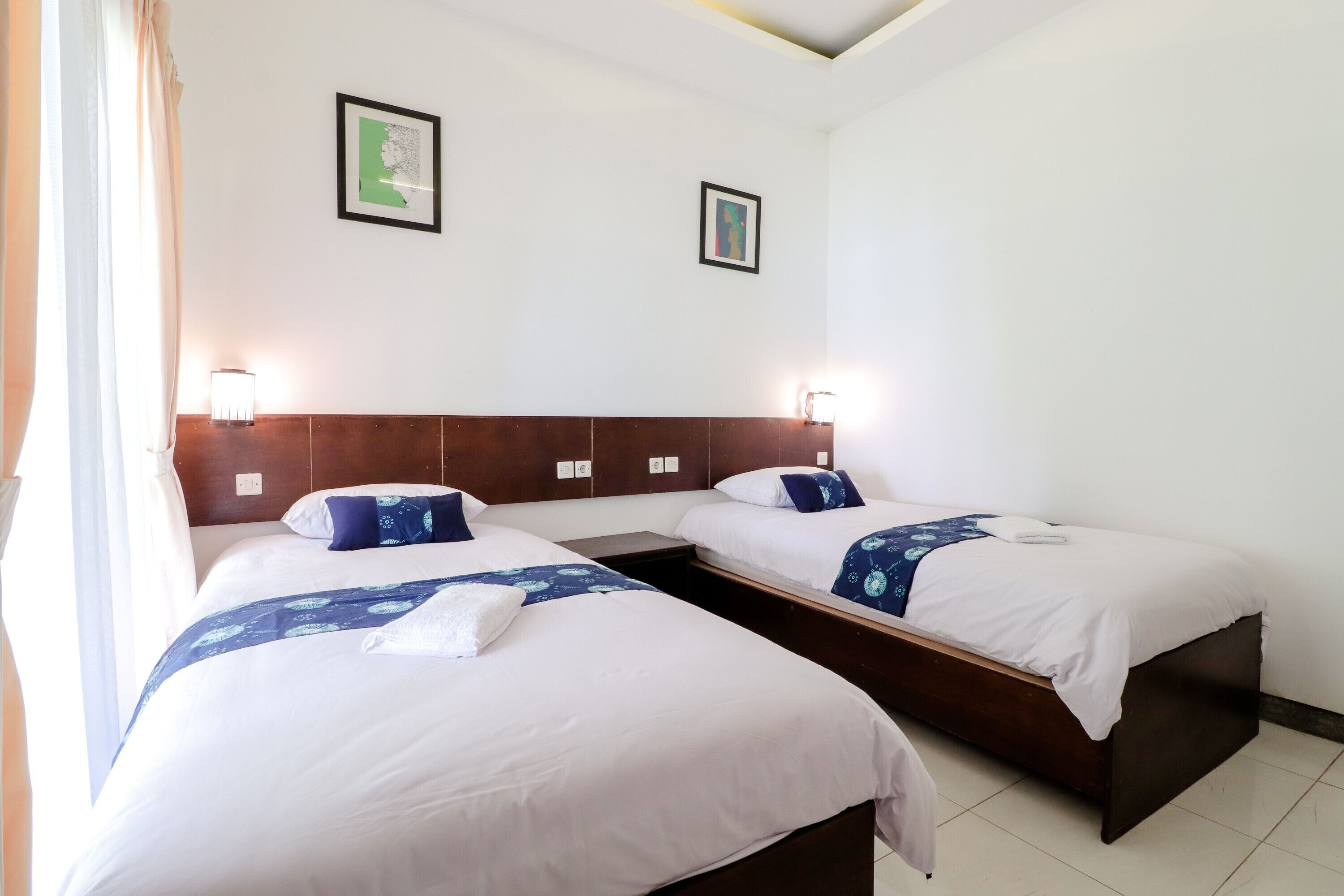 Una habitación de hotel con dos camas individuales, cada una con cabecero, ropa de cama blanca y una manta azul estampada. Hay dos lámparas de pared a cada lado de las camas, y un mapa enmarcado en la pared.
