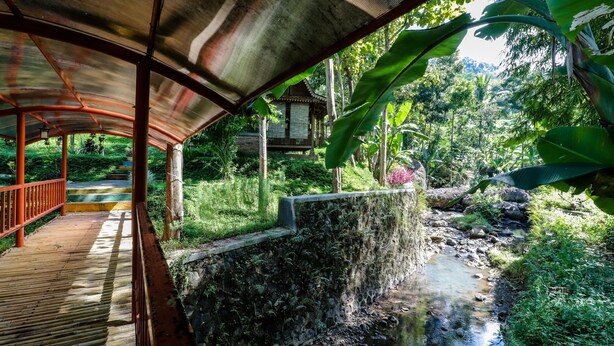 A covered wooden walkway leading to a traditional house with a stone wall and a stream. - Balkondes Kenalan (Borobudur)