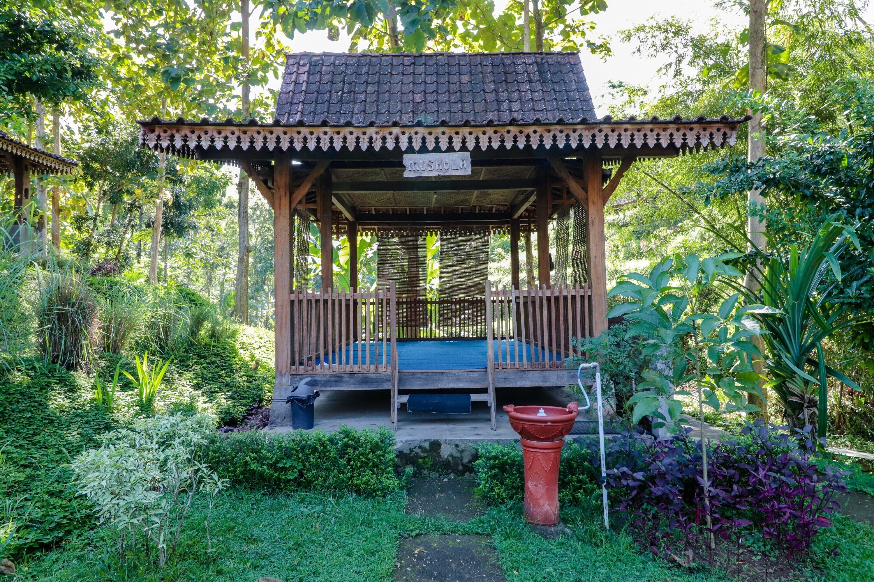 A wooden gazebo with a tiled roof, surrounded by lush greenery and a small water feature.