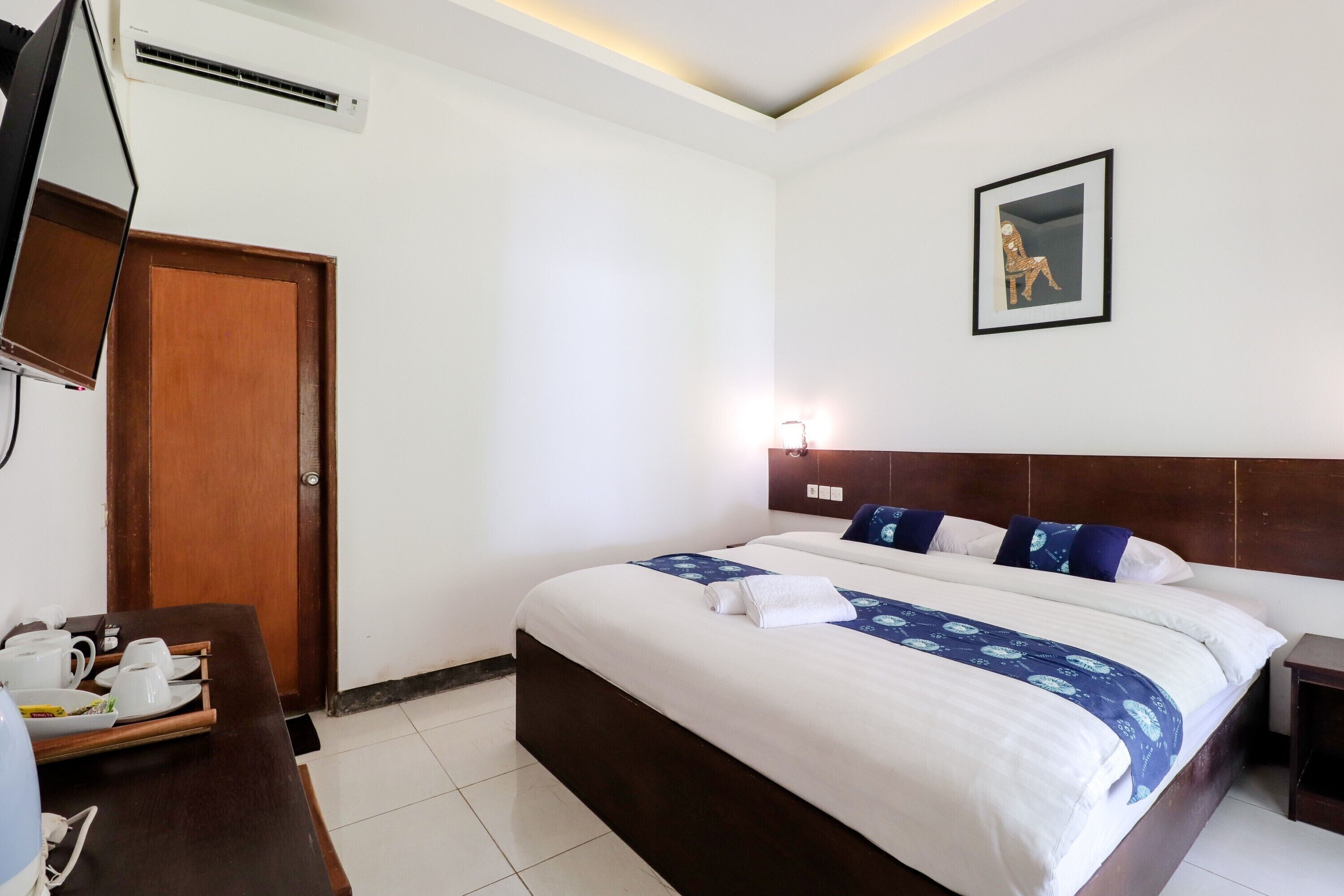 A hotel room with a bed, a bedside table, a desk with a chair, a television, a door, and a wall-mounted picture.