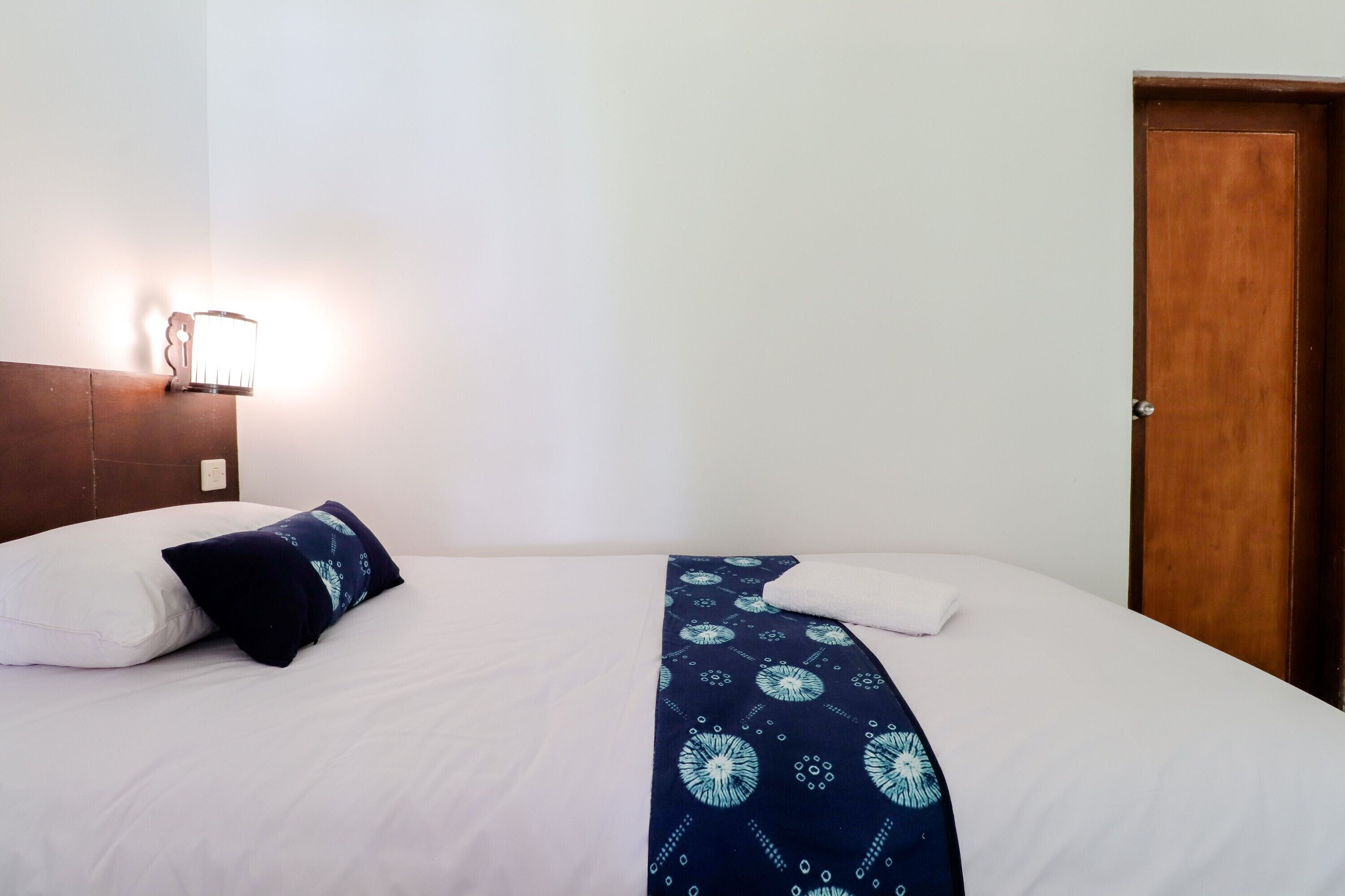 Una cama bien hecha con ropa de cama blanca y azul oscuro, una lámpara fijada a la pared y una puerta de madera.