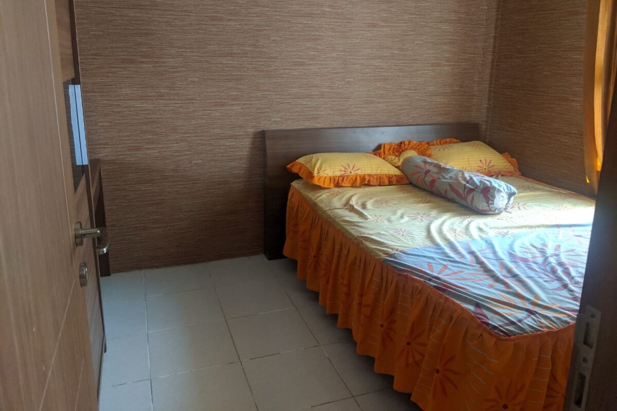 Un petit lit bien fait, avec une couvre-lit orange et des oreillers, une tête de lit en bois et un sol carrelé.